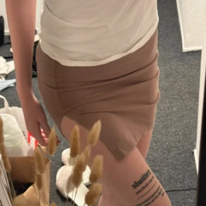 Beige kortkjol med omlott-detalj - Säljer en beige kortkjol med omlott-detalj och lätt asymmetrisk nederkant. Kjolen har en tight passform och är tillverkad i ett mjukt, stretchigt material som sitter snyggt på kroppen. Perfekt för dig som gillar stilrena och enkla plagg.