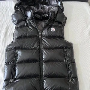 Moncler väst - Säljer en svart, glansig dunväst från Moncler med huva och två dragkedjefickor framtill. Västen har quiltad design och stängs med dubbel dragkedja. Klassisk Moncler-logga på bröstet och serietidningsdetalj på insidan.