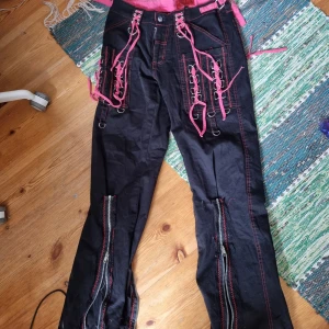Tripp -liknande byxor - Tipp-liknande svarta byxor med rosa snörning och silverfärgade ringdetaljer framtill. Byxorna har dekorativa dragkedjor längs benen och kontrastsömmar i rosa. Lågmidjade och utsvängda. Perfekt för en emo look jag hade 100% sparat dem om jag fortfarande passade i dem. 