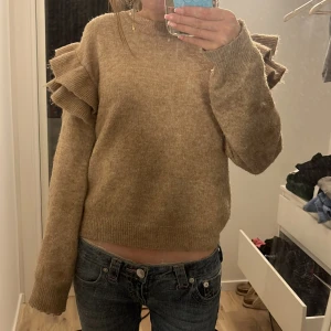 Beige stickad tröja med volangdetaljer - Säljer en beige stickad tröja med långa ärmar och volangdetaljer på axlarna. Tröjan har en rund halsringning och är mjuk och skön. Perfekt för dig som gillar unika detaljer och en mysig stil.