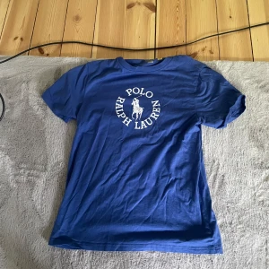 Blå t-shirt från Polo Ralph Lauren - Säljer en blå t-shirt från Polo Ralph Lauren med klassisk vit logga och text på bröstet. T-shirten har rund hals och korta ärmar. Perfekt för en avslappnad stil.