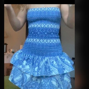 Blå och vit axelbandslös kortklänning med mönster - Säljer en söt axelbandslös kortklänning i blått och vitt med smock upptill och volangkjol. Klänningen har ett detaljerat mönster och passar perfekt för varma dagar.