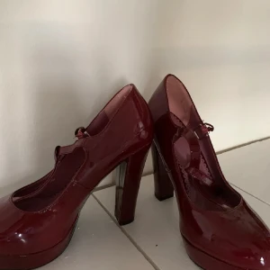 Vinröda lackpumps med rem - Säljer ett par vinröda pumps i glansigt lackmaterial med rund tå och hög klack. Skorna har en smal rem över foten som ger en klassisk Mary Jane-look. Perfekta för dig som vill sticka ut med en elegant och trendig stil.