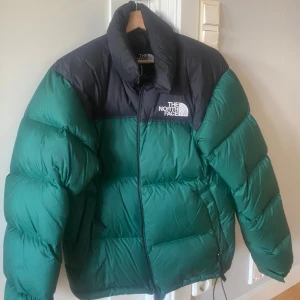 Grön och svart pufferjacka från The North Face - Säljer en grön och svart pufferjacka från The North Face. Jackan har lång ärm, dragkedja och klassisk logga på bröstet och ryggen. Perfekt för kyliga dagar med sin tjocka och vadderade design.