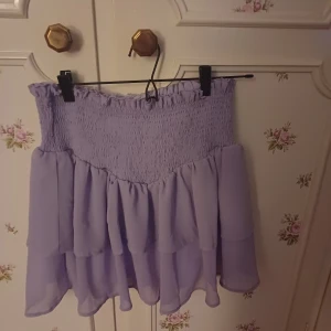 Ljuslila volangkjol - Supersöt ljuslila kjol med smock i midjan och dubbla volanger. Kjolen är kort och har en flowy känsla, perfekt för varma dagar. Passar dig som gillar en feminin och trendig stil.