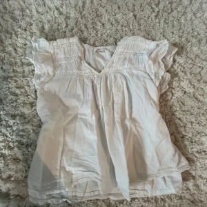 Vit blus med volangärmar från Gina Tricot - Säljer en söt vit blus från Gina Tricot med volangärmar och fina broderade detaljer upptill. Blusen har v-ringning och är tillverkad i ett lätt bomullsmaterial. Perfekt för vår och sommar!