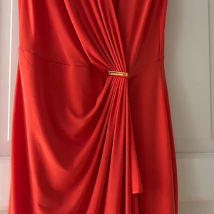 Orange omlottklänning från Michael Kors - Säljer en snygg röd omlottklänning från Michael Kors med guldfärgad detalj i midjan. Klänningen har draperad front och är ärmlös, perfekt för dig som vill sticka ut med en elegant look.