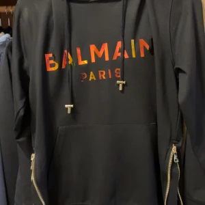 Svart hoodie från Balmain Paris med färgglad logga - Säljer en svart hoodie från Balmain Paris med en färgglad logga framtill. Tröjan har huva med dragsko, magficka och dragkedjor i sidorna. Perfekt för dig som gillar streetwear och vill sticka ut med en exklusiv look.