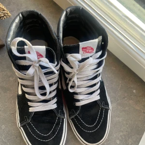 Svarta höga sneakers från Vans - Säljer ett par klassiska svarta Vans sneakers med vit sula och vit sidorand. Skorna har högt skaft och vita snören, perfekt för en streetwear-look. Ikonisk design med rund tå och platt sula.