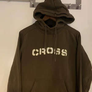 Brun hoodie från Cross - Säljer en brun hoodie från Cross med vit text på bröstet. Tröjan har huva med dragsko och en stor magficka framtill. Perfekt för en avslappnad stil.