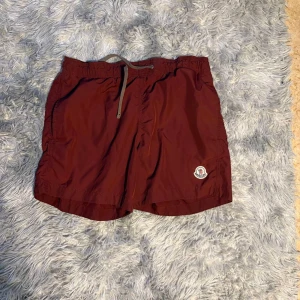 Vinröda badshorts från Moncler - Snygga vinröda badshorts från Moncler med snörning i midjan och klassisk Moncler-logga på benet. Shortsen har innerbyxa i mesh och är perfekta för sommardagar vid stranden eller poolen.