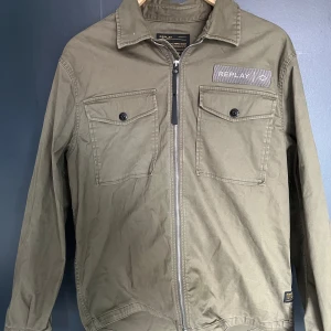 Olivgrön overshirt från Replay - Snygg olivgrön overshirt från Replay med dragkedja framtill och två bröstfickor med knappar. Märkespatch på vänster bröst och klassisk krage. Perfekt för lager-på-lager och stilren look.