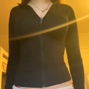 Svart tight zip up - Säljer en svart, tight zip up med dragkedja framtill. Den  har lång ärm och en kroppsnära passform som framhäver siluetten. Perfekt att slänga på sig över en topp eller sport-bh. Enkel och stilren design utan synliga loggor.