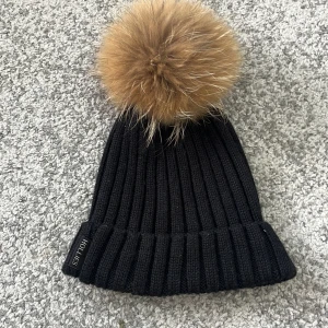 Svart ribbad mössa med äkta pälsboll från Hollies - Snygg svart ribbstickad mössa från Hollies med stor äkta pälsboll på toppen. Perfekt accessoar för kyliga dagar och ger en trendig look. Mössan har uppvikt kant och diskret logga på sidan.