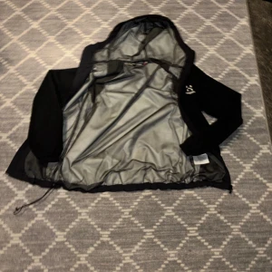 Svart vindjacka från Haglöfs med huva - Säljer en svart vindjacka från Haglöfs i storlek M. Jackan har huva, dragkedja framtill och justerbar nederkant med dragsko. Insidan är fodrad med mesh för extra komfort. Perfekt för blåsiga dagar.