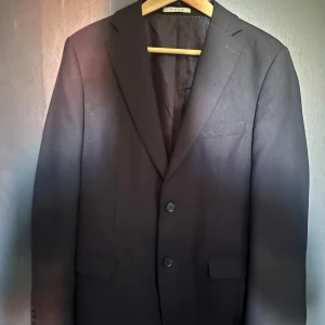 Svart kavaj från POUR - Stilren svart kavaj från POUR med klassisk passform och två knappar framtill. Kavajen har två fickor med lock och en bröstficka. Perfekt för formella tillfällen och enkel att matcha med skjorta eller t-shirt.