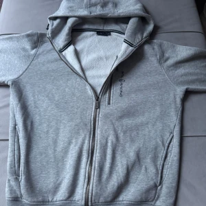 Grå hoodie från Sail Racing - Säljer en ljusgrå hoodie från Sail Racing med dragkedja framtill och en extra ficka med dragkedja på bröstet. Tröjan har huva och är tillverkad i mjukt material, perfekt för en avslappnad stil.