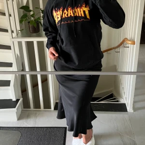 Svart Thrasher hoodie med flames-logga - Säljer en svart hoodie från Thrasher med den klassiska flames-loggan i gult och rött på bröstet. Tröjan har huva med dragsko och en stor magficka. Perfekt för dig som gillar streetwear och skate-stil🛹🔥❤️‍🔥