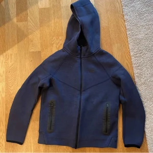 Mörkblå hoodiejacka från Nike - Mörkblå hoodiejacka från Nike med dragkedja framtill och två fickor med dragkedja. Jackan har huva och är långärmad, perfekt för svala dagar. Nike-logga på bröstet. Snygg och sportig stil.