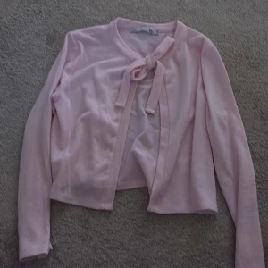 Ljusrosa kofta med rosett - Söt ljusrosa kofta från lager 157 i storlek XS. Koftan har långa ärmar och en fin rosettdetalj framtill vid halsen. Perfekt att slänga över en topp för en gullig look.