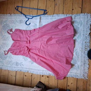 Rosa axelbandsklänning med volangdetaljer - Söt rosa klänning med axelband och volangdetaljer upptill. Klänningen har en utställd kjol och knytning i midjan för en feminin siluett. Perfekt för sommarens alla tillfällen.