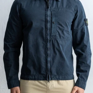 Mörkblå overshirt från Stone Island - Säljer en mörkblå overshirt från Stone Island med dragkedja och knappstängd bröstficka. Klassisk patch på vänster ärm. Perfekt för lager-på-lager och stilren look. Den är i använt skick och har ett hål på ena armbågen. Just denna modell säljs inte längre men liknande kostar nu 4800kr