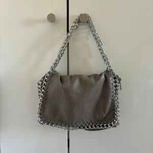 Tiamo väska  - Grå och tiamo väska i mellan storlek. Finns en lång kedja som man även kan vika så att det blir ett kortare band om det önskas.   Den är använd en del men kommer inte längre till användning så säljer därför den⭐️ Den har några små slitage, på insidan är den lite svart (se bild), och har tappat lite färg på vissa ställen.   Tror jag köpte den på scorett för ca 450 kr.