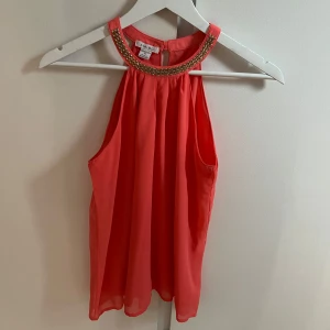 Korallfärgad ärmlös topp  - Säljer en korallfärgad ärmlös topp från Amisu med snygg guldfärgad kedjedetalj vid halsringningen. Toppen har en lös passform och knäpps i nacken. Perfekt för att lyfta en outfit med lite extra bling!