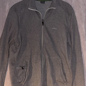 Grå zip-tröja från BOSS - Säljer en stilren grå zip tröja från BOSS med dragkedja framtill och diskret logga på bröstet. Jackan har långa ärmar, ståkrage och två fickor med dragkedja. Perfekt för lager-på-lager och enkel att matcha.