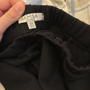 Svarta byxor från Mamalicious - Svarta byxor från Mamalicious med elastisk midja och lös passform. Tillverkade i ett mjukt och lätt syntetmaterial som ger en skön känsla. Perfekta för dig som vill ha bekväma och stilrena byxor.
