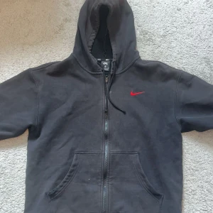 Vintage Svart hoodie från Nike SB - Svart hoodie från Nike SB med dragkedja framtill och röd Nike-logga på bröstet. Tröjan har huva och två stora fickor framtill. Perfekt för en avslappnad stil.