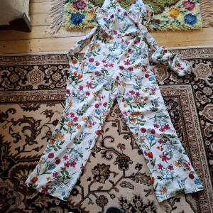 Vit blommig byxdress med knytband - Säljer en vit byxdress med färgglatt blommönster och smala axelband. Byxdressen har knytband i midjan och vida ben. Perfekt för varma dagar och ger en somrig känsla.