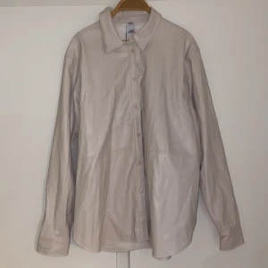 Beige skinnskjorta - Säljer en beige skjorta i skinnimitation med klassisk krage och knäppning framtill. Skjortan har långa ärmar och en rak passform, perfekt för en stilren look.