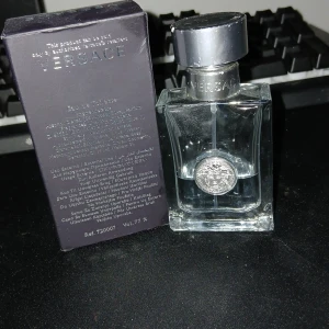 Versace Pour Homme Eau de Toilette - Versace Pour Homme Eau de Toilette i en elegant, genomskinlig glasflaska med silverfärgat lock och Versace-logga framtill. Parfymen kommer i en stilren mörklila kartong med silverdetaljer. En klassisk och fräsch doft för herr.
