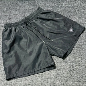 Svarta shorts från Prada med snörning - Svarta shorts från Prada med elastisk midja och snörning. Klassisk design med diskret Prada-logga på vänster ben. Tillverkade i ett lätt och glansigt syntetmaterial med meshfoder för extra komfort.