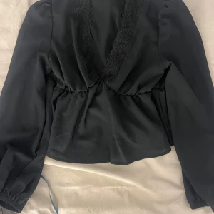 Svart blus med spetsdetalj - Säljer en svart blus med djup v-ringning och spetsdetaljer längs ringningen. Blusen har långa ärmar med lätt puff och resår vid ärmslut. Modellen är något croppad och har en feminin siluett.