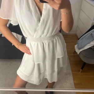 Vit omlottklänning med volang - Säljer en söt vit kortklänning med omlottliv och volangdetaljer. Klänningen har resår i midjan och är ärmlös med lätt v-ringning. Perfekt för sommarens tillfällen!