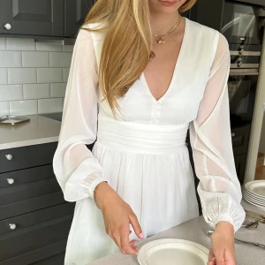 Vit klänning med v-ringning och långa ärmar - Säljer en vit klänning med v-ringning, långa transparenta ärmar och markerad midja med knytband i ryggen. Kjolen är lätt utställd och klänningen har en luftig känsla. Perfekt för skolavslutning eller sommarfest. Klänningen är från Bubbelroom. Stl 32, för mått skicka dm.. 