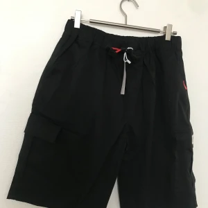 Svarta cargoshorts med snörning - Svarta cargoshorts med elastisk midja och vit snörning framtill. Shortsen har flera fickor på sidorna och är perfekta för en avslappnad stil. Passar bra till sommaren och har en normal passform. Stlk kvar M-4XL 