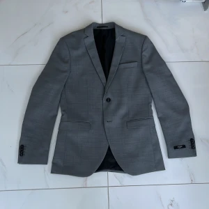 Grå kostym med kavaj och byxor - Snygg grå kostym med klassisk kavaj och matchande byxor. Kavajen har två knappar, smal passform och fickor framtill. Perfekt för bal eller formella tillfällen. Tidlös design i ett lätt material. Använd endast en gång och är så gott som ny. Slim fit passform. Kommer i tillhörande kostymbag!