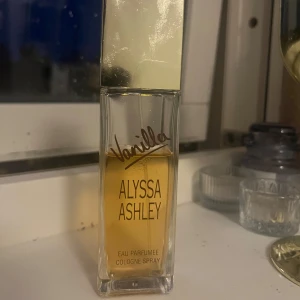 Vanilla Eau Parfumée Cologne Spray från Alyssa Ashley - En parfym med doft av vanilj från Alyssa Ashley. Flaskan är rektangulär i genomskinligt glas med guldig kork och text i guld. Innehållet är ljusgult och flaskan har en stilren design.