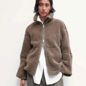 Brun teddyjacka med dragkedja - Mysig brun teddyjacka med hög krage och hel dragkedja framtill. Jackan har två stora fickor och en oversized passform som ger en avslappnad look. Perfekt för kyliga dagar och enkel att styla över en skjorta eller tröja.