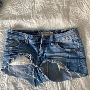 Ljusblå jeansshorts med slitningar - Säljer ett par ljusblå jeansshorts med kraftiga slitningar och hål framtill. Klassisk femficksmodell, låg midja och råa kanter nertill. Perfekta för sommaren och en avslappnad stil.