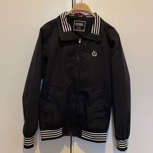 Svart vindjacka från Etirel med vita ränder - Svart vindjacka från Etirel med vita ränder på krage, ärmslut och midja. Jackan har dragkedja framtill och broderad FC-logga på bröstet. Klassisk sportig look med normal passform. En jacka som funkar för stilen och mot dålig väder 😃