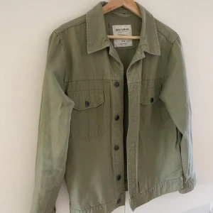 Olivgrön jeansjacka från Pull&Bear - Säljer en olivgrön jeansjacka från Pull&Bear i storlek S. Jackan har klassisk krage, knäppning framtill och två bröstfickor med lock och knapp. Perfekt för lager-på-lager och enkel att styla till olika outfits.