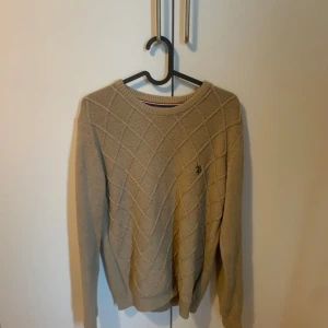 Beige stickad tröja med rutmönster - Säljer en beige stickad tröja från Polo assn med diskret rutmönster och rund halsringning. Tröjan har långa ärmar och en liten broderad logga på bröstet. Perfekt för lager-på-lager under kyliga dagar.