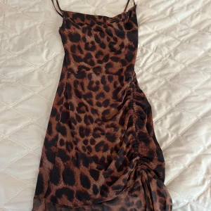 Leopardmönstrad kortklänning från PrettyLittleThing - Säljer en snygg leopardmönstrad kortklänning från PrettyLittleThing. Klänningen har smala axelband, åtsittande passform och rynkade detaljer på sidan. Perfekt för dig som vill sticka ut med ett djurmönster och trendig look.