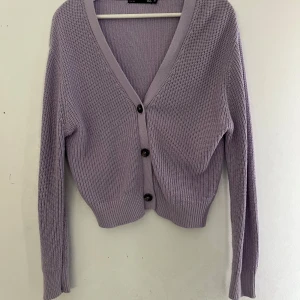 Ljuslila stickad kofta med knappar - Säljer en söt ljuslila stickad kofta med v-ringning och tre knappar framtill. Koftan har lång ärm och ribbade muddar vid ärmslut och nederkant.