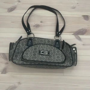 Grå axelväska med monogrammönster - Säljer en snygg grå axelväska som liknar  Guess eller är guess är inte helt säker med svart monogrammönster och svarta detaljer. Väskan har dubbla handtag, dragkedja upptill och flera fack. 