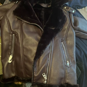 Mörk lila med lite brun skinnjacka med fuskpäls från Joinus - Säljer en brun skinnjacka från Joinus med mjuk mörk fuskpäls på krage och insida. Jackan har silvriga dragkedjor, två snedställda fickor och markerade sömmar. Perfekt för kyliga dagar och ger en cool look.